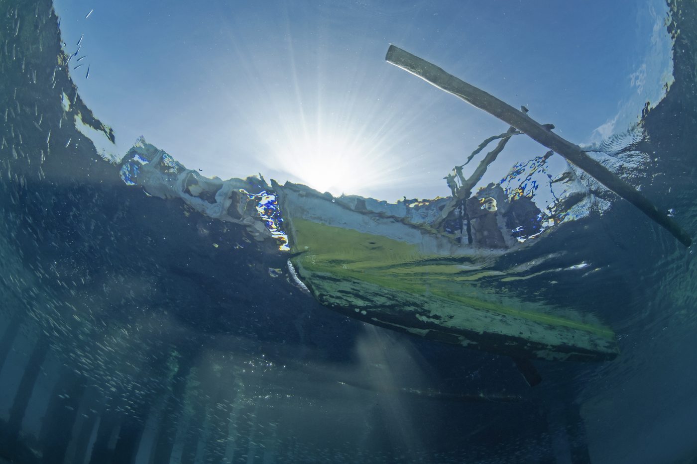Sunlight bursting through the water's surface beneath the hull of an outrigger canoe.