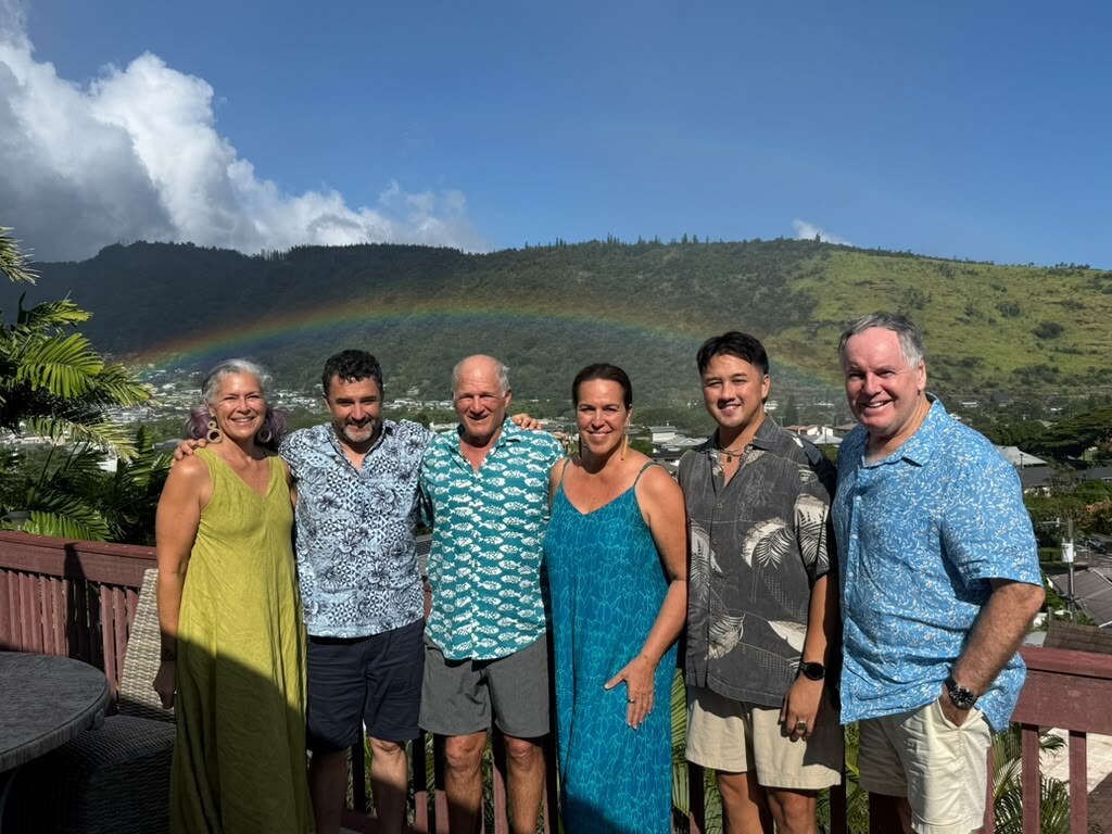 Members of Big Ocean's board and operations team standing together on a lanai, a rainbow arching over the mountains behind them.